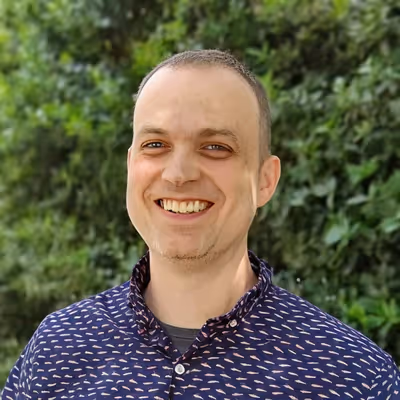 Smiling man with short hair wearing a patterned navy blue shirt standing in front of green foliage.