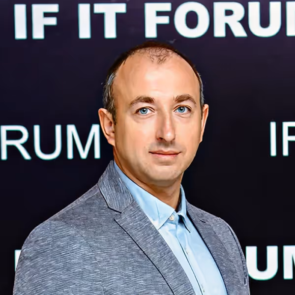 Man with blue eyes wearing a light blue shirt and gray blazer, standing in front of a black background with partially visible white text.