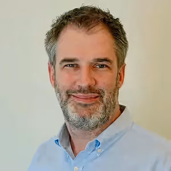 Smiling middle-aged man with salt-and-pepper hair and beard wearing a light blue button-up shirt.
