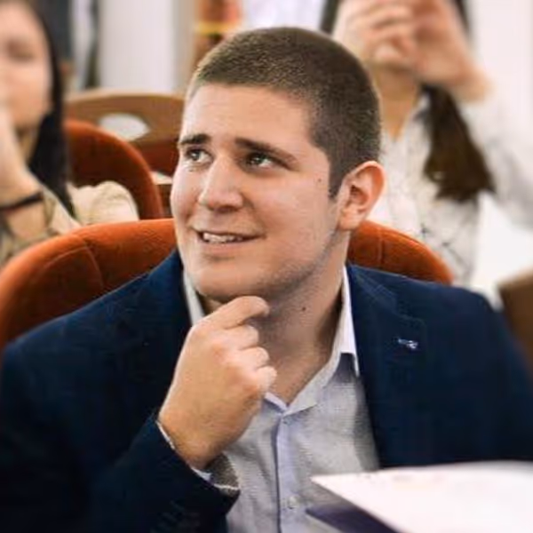 Young man in a navy blazer sitting in a chair, looking to the side with a thoughtful expression and hand near his chin.