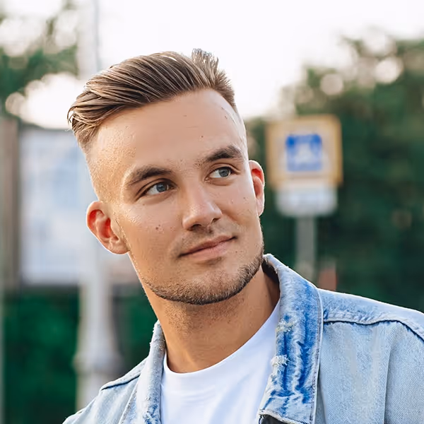 Young man with styled blonde hair wearing a light denim jacket and white shirt, looking slightly to the side outdoors.